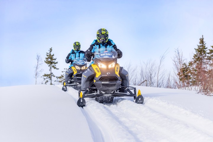 Two people in helmets riding snowmobiles on a snowy trail.