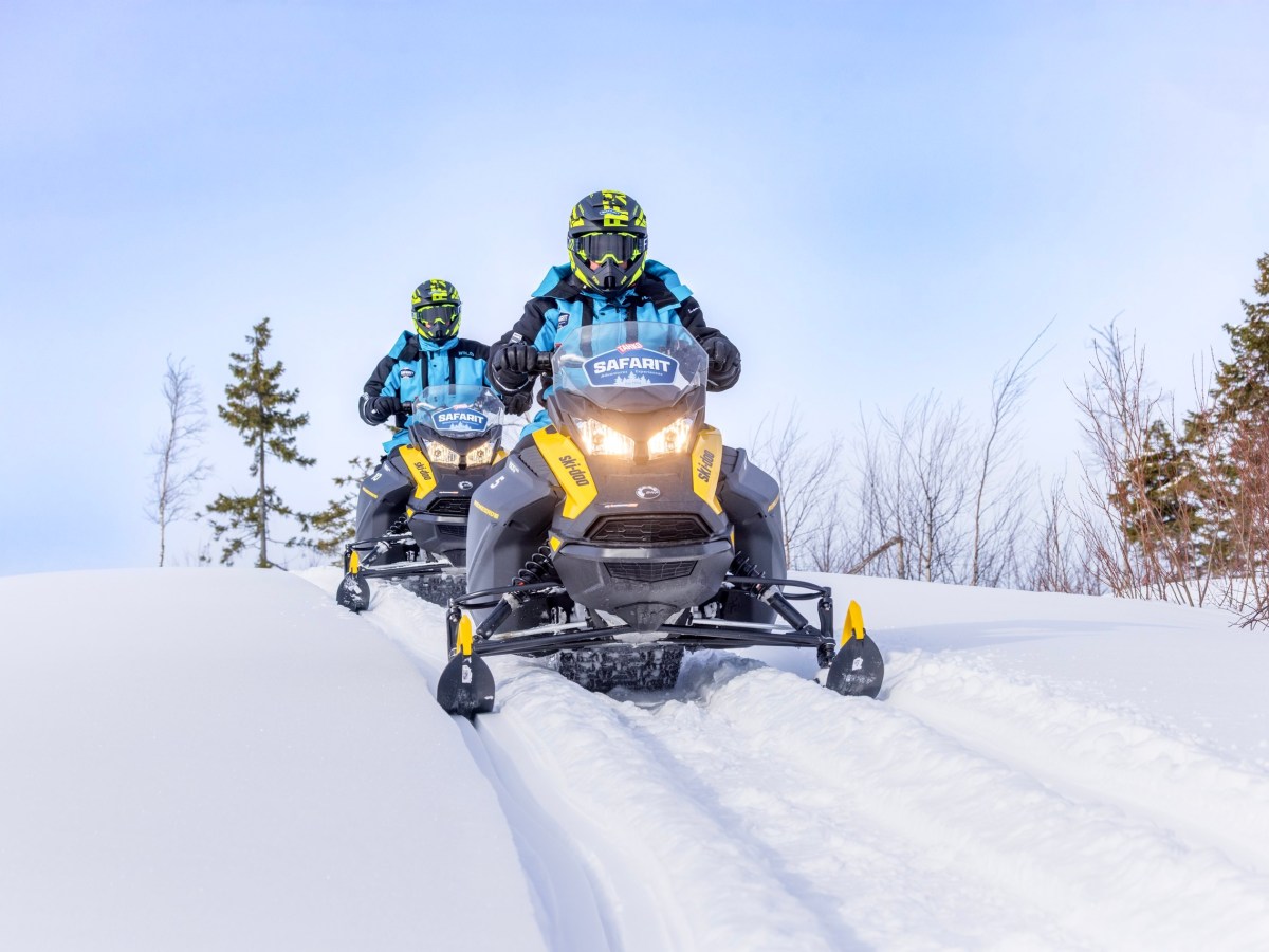 Two people in helmets riding snowmobiles on a snowy trail.