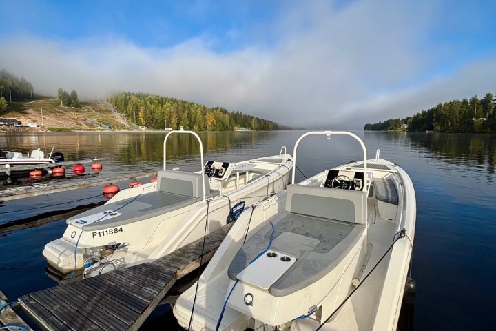 Two white boats docked on a calm lake under a blue sky with trees in the background.