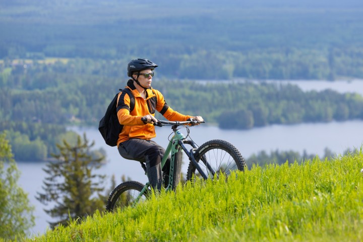 Person in orange jacket mountain biking on a grassy hill with a lake and trees in the background.