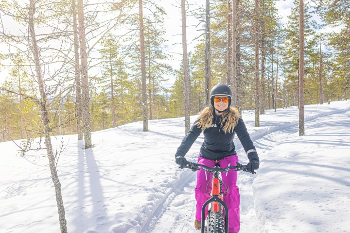 Person cycling on a snowy forest trail with tall trees and bright sunlight.