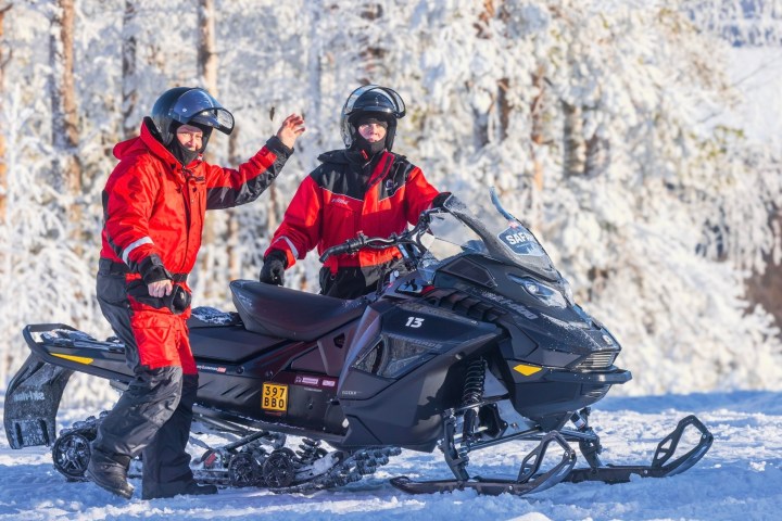 Two people in red outfits with helmets stand next to a snowmobile in a snowy landscape.
