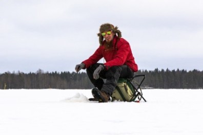 Person ice fishing on a frozen lake, wearing a red jacket and furry hat.