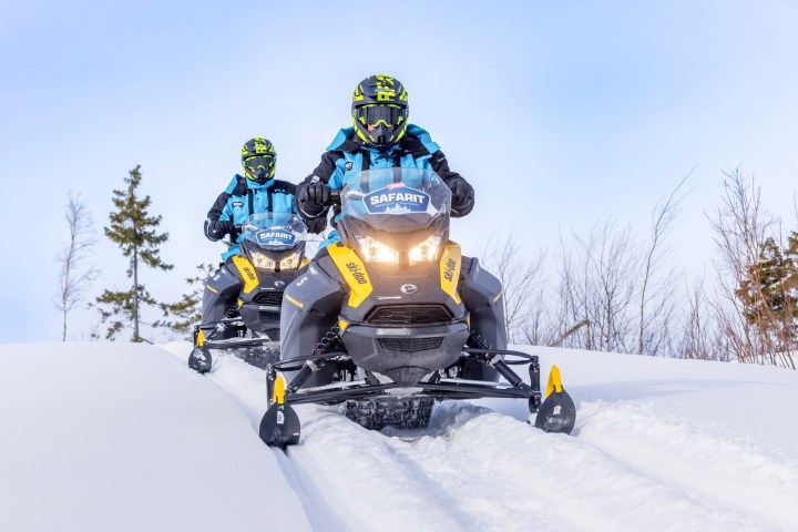 Two people snowmobiling on a snowy trail with trees in the background.