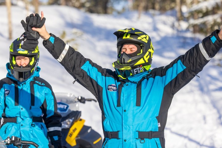 Two people in blue snow gear celebrate on snowmobiles in snowy forest.