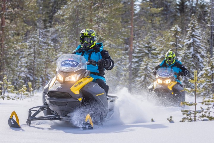 Two people riding snowmobiles through a snowy forest landscape.