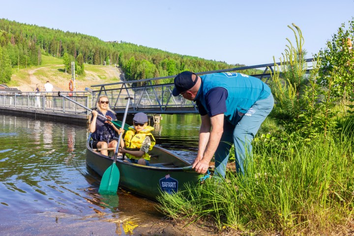 Adult and child in life vests canoeing on a lake, with man assisting from the shore.