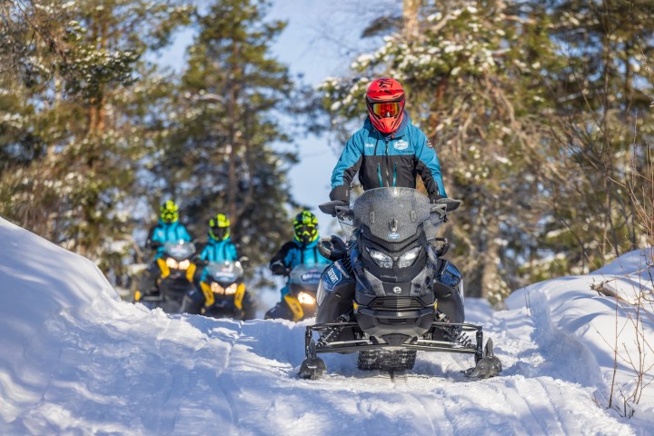 Four people riding snowmobiles on a snowy trail through a forest.