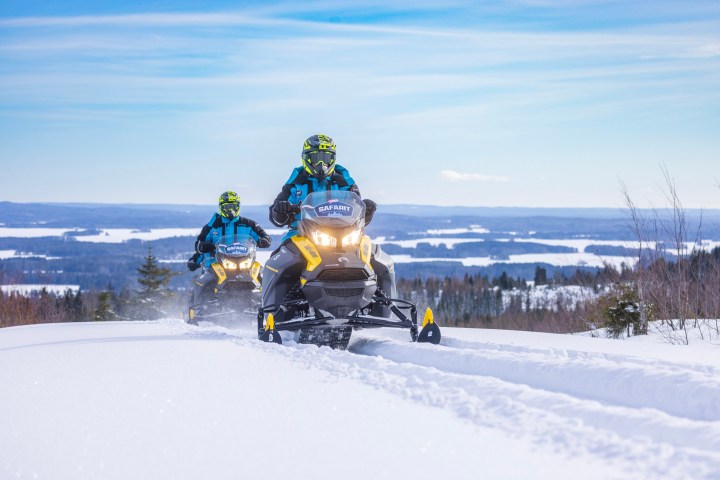 Two people riding snowmobiles on a snowy hill under a clear blue sky.