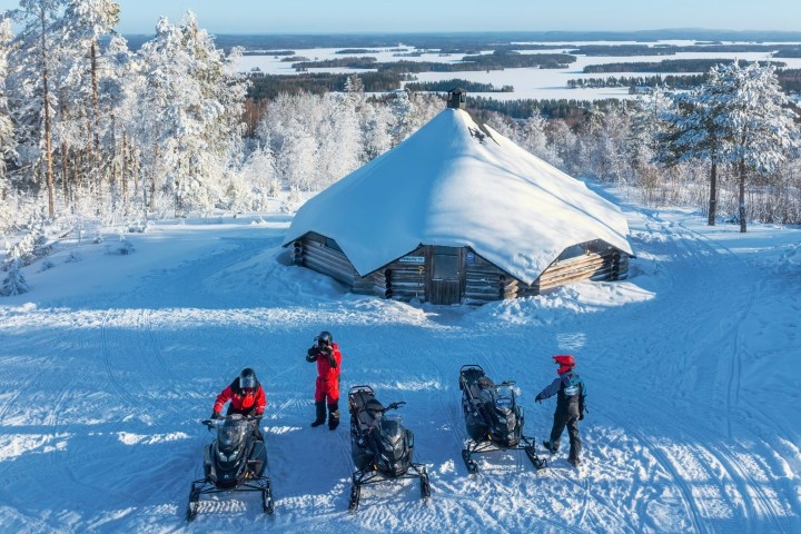 People on snowmobiles outside a snowy cabin in a winter landscape.