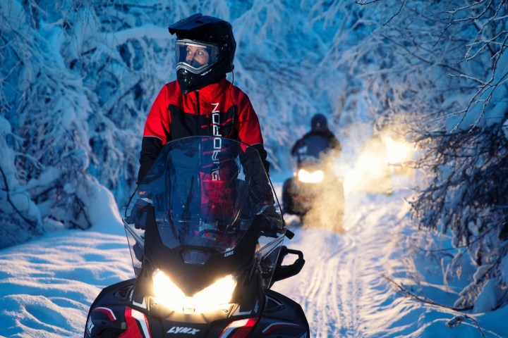 Two snowmobilers ride through a snowy forest trail.