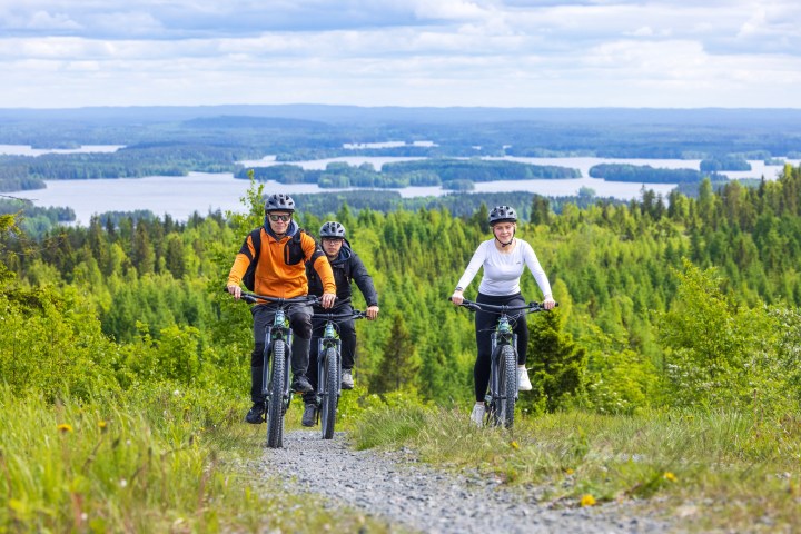 Three cyclists on a trail with forest and lake view in the background.