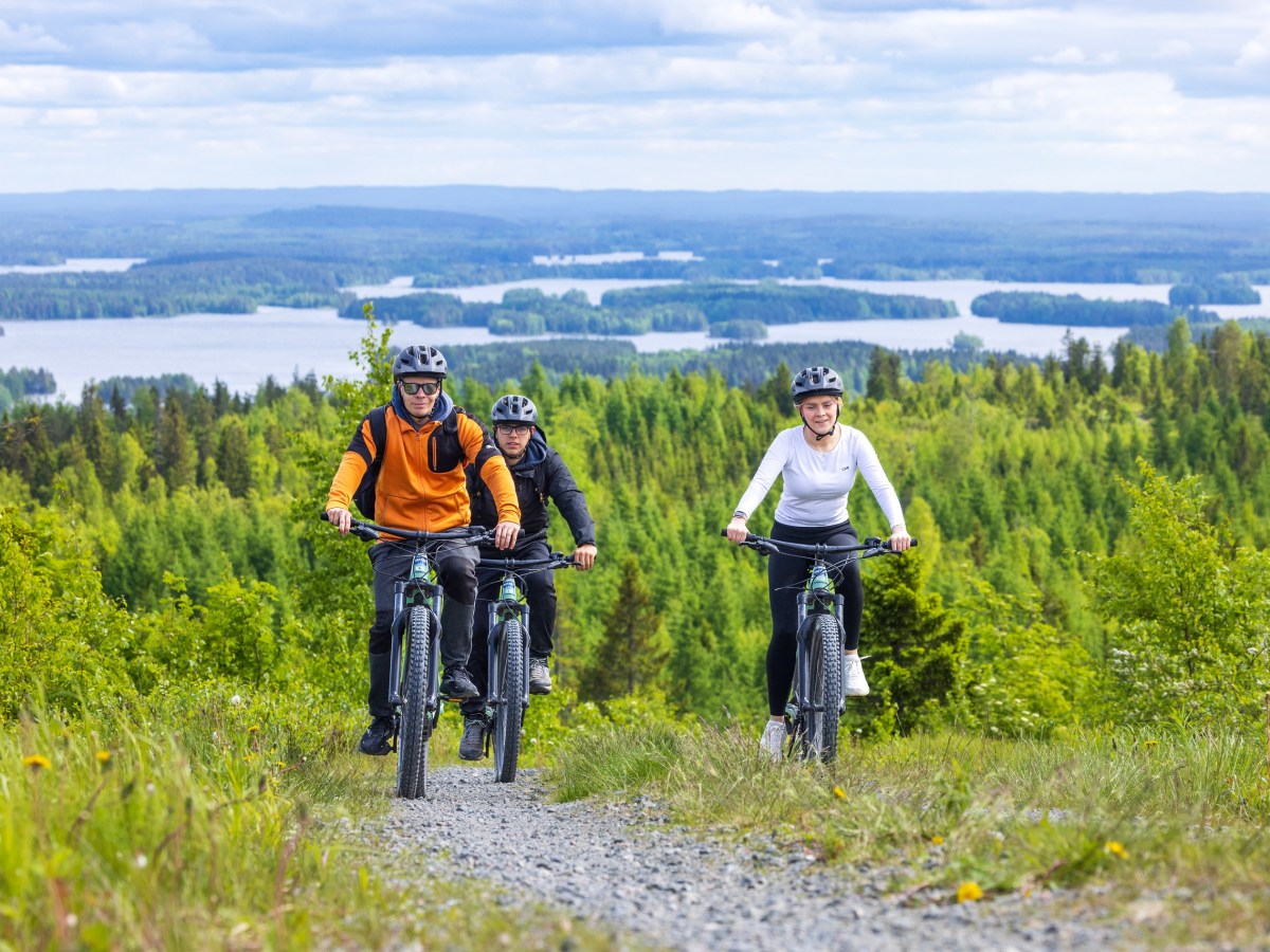 Three cyclists on a trail with forest and lake view in the background.