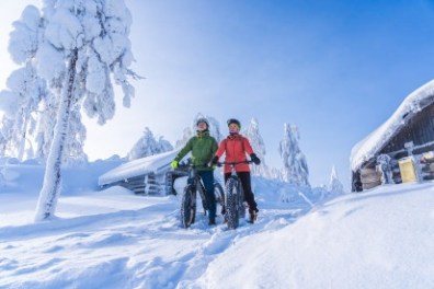 Two people biking on a snowy trail near cabins and snow-covered trees.