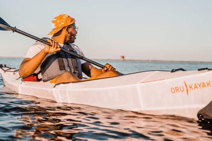 Man kayaking on calm water in a foldable kayak, wearing a life vest and hat.