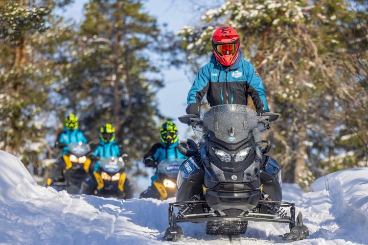 Four people riding snowmobiles on a snowy forest trail.