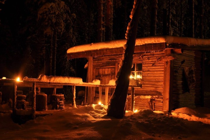 Cozy wooden cabin in snowy woods at night, lit by warm lanterns.
