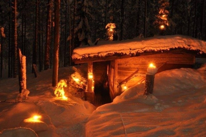 Snow-covered cabin in forest illuminated by warm lights at night.