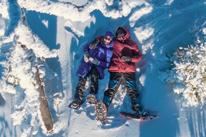 Two people in winter gear lying on snow among trees, viewed from above.