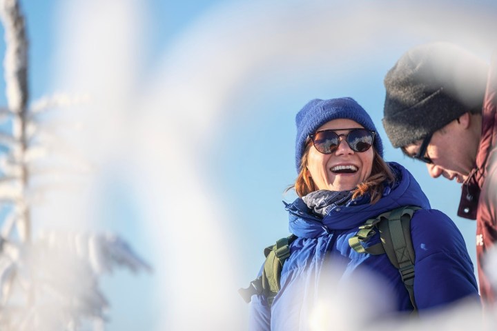 Two people in winter clothing outdoors, one smiling, snow-covered tree in foreground.