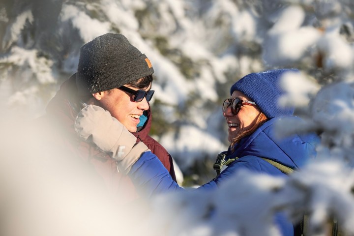 Two people in winter clothing smiling amidst snow-covered trees.