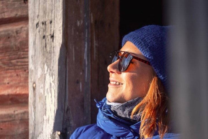 Woman in blue beanie and sunglasses smiling by a wooden wall.