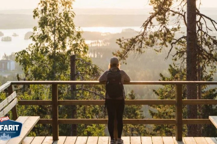 Person with backpack stands on a wooden deck overlooking a forest and lake at sunset.