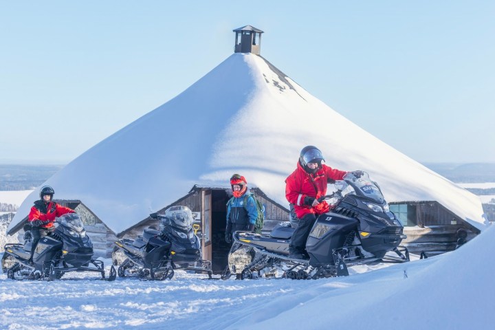 People on snowmobiles in front of a snow-covered cabin.
