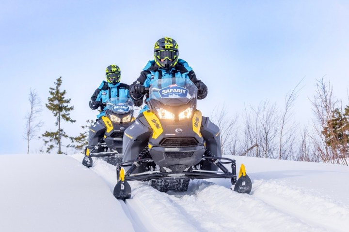 Two people riding snowmobiles on a snowy trail with trees in the background.