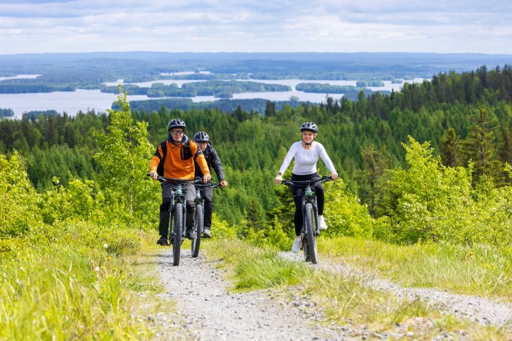 Three cyclists riding on a gravel path through a lush forest with a lake in the background.