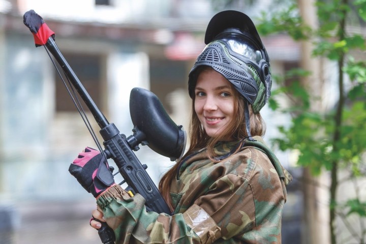 Person in camo gear holding a paintball gun, smiling outdoors.