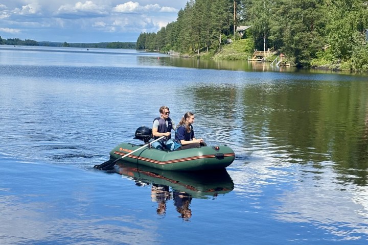 Two people in an inflatable boat on a calm lake with trees in the background.