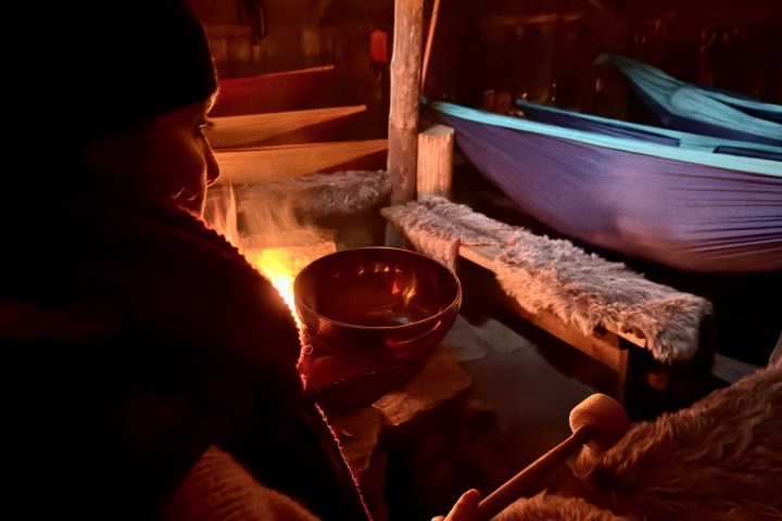 Person holding bowl by fire in dimly lit rustic cabin with hammocks and fur-covered benches.