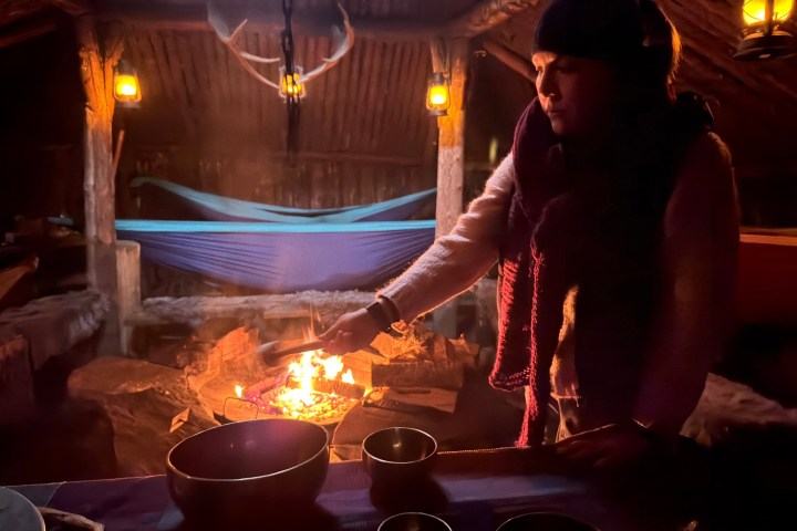 Person tending fire in cozy, dimly-lit cabin with hammock, bowls, and rustic décor.