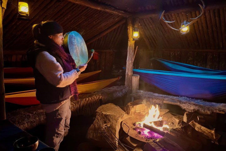 Person playing a drum by a campfire in a rustic cabin with hammocks and lanterns.